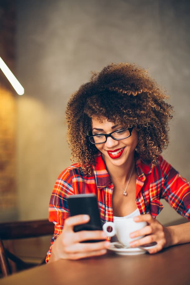 Une femme est assise à une table avec une tasse de café et sourit à son téléphone en regardant TriFact365, le logiciel de traitement des factures numériques.