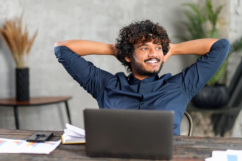 Man with his arms behind his head sits relaxed behind his laptop