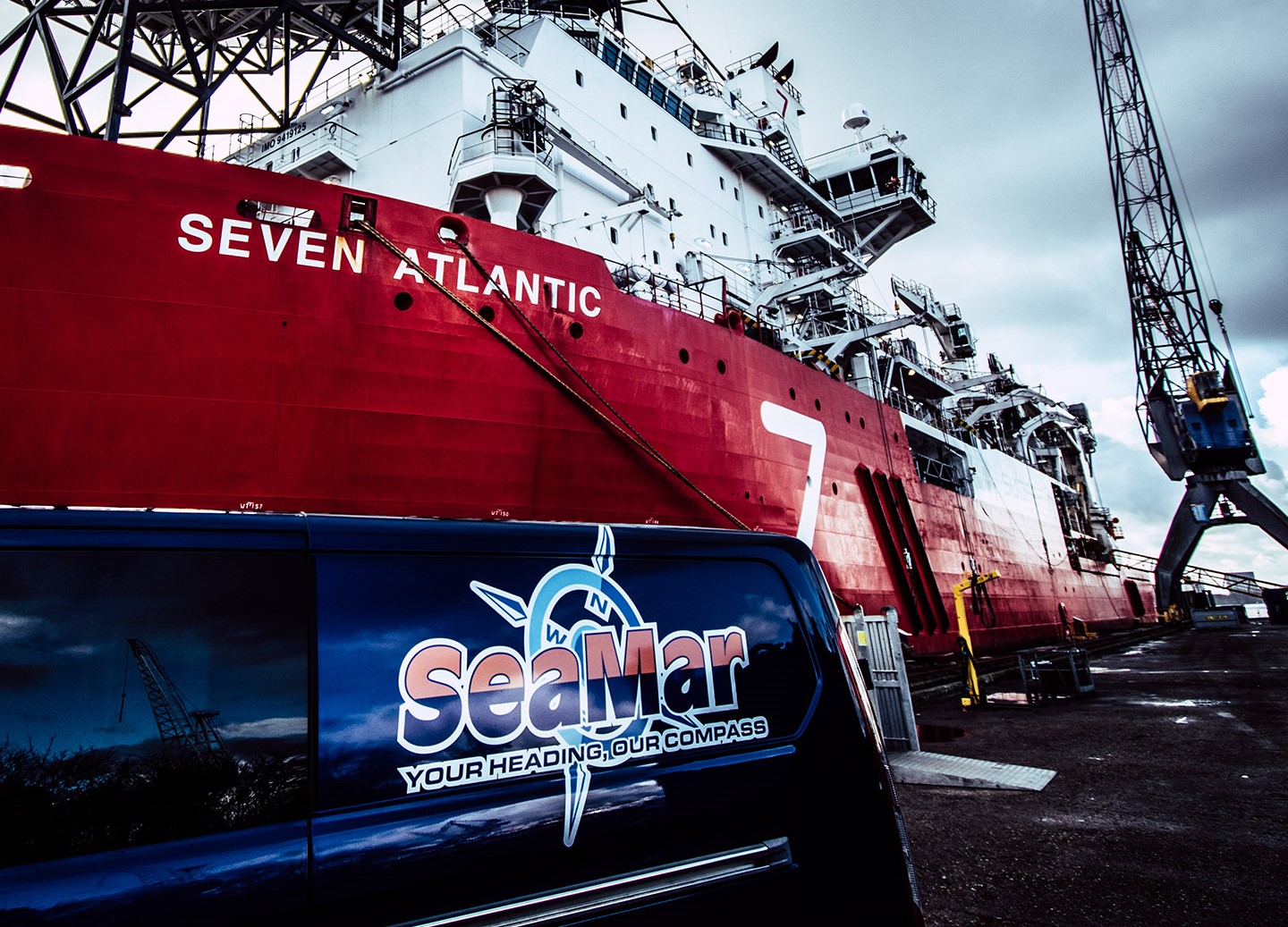 Een busje van SeaMar in de haven met op de achtergrond het schip "Seven Atlantic" en een kraan