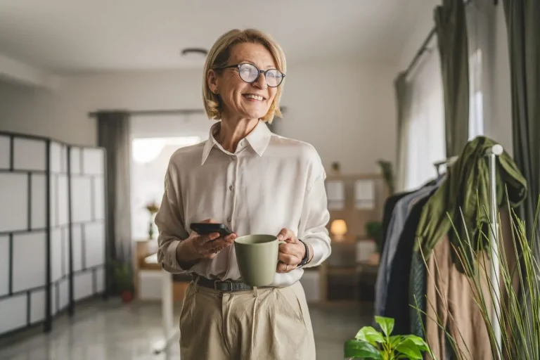 Vrouw kijkt lachend naar buiten, terwijl ze in de ene hand een kop koffie heeft en in haar andere hand een mobiele telefoon.