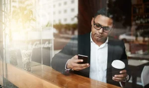 Man met een bril op en een koffiekop in zijn linkerhand kijkt op zijn telefoon in zijn rechterhand.