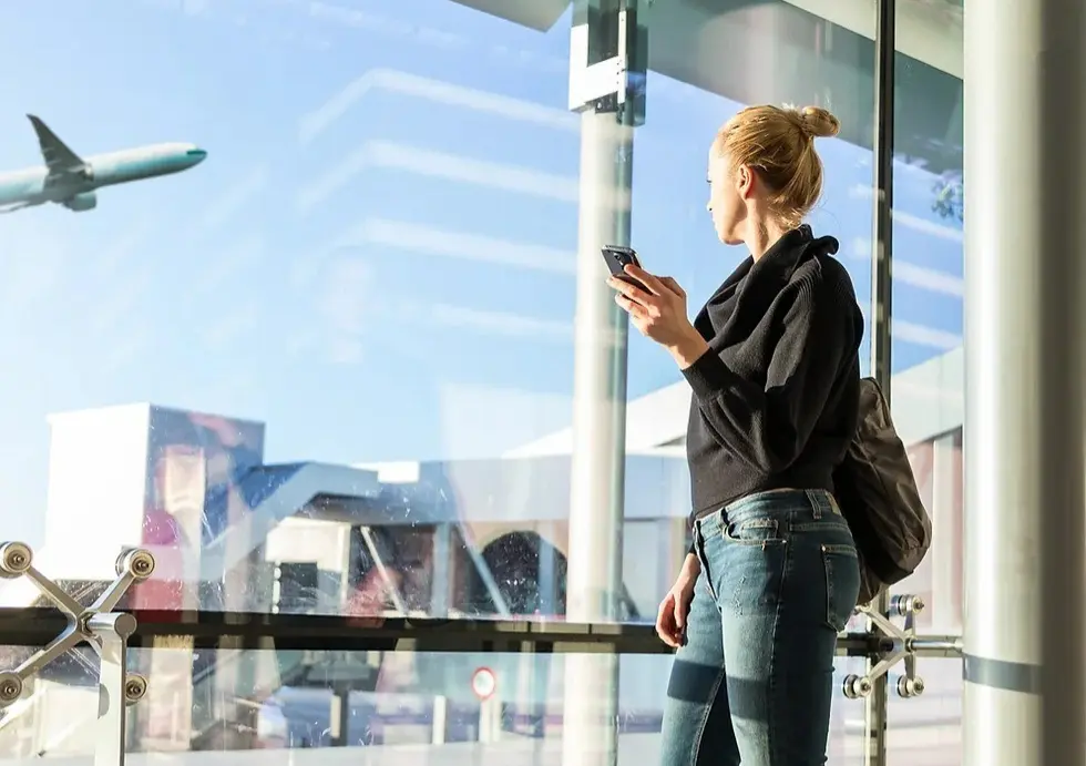 Vrouw kijkt op een vliegveld uit het raam, terwijl ze haar telefoon in haar hand houdt.