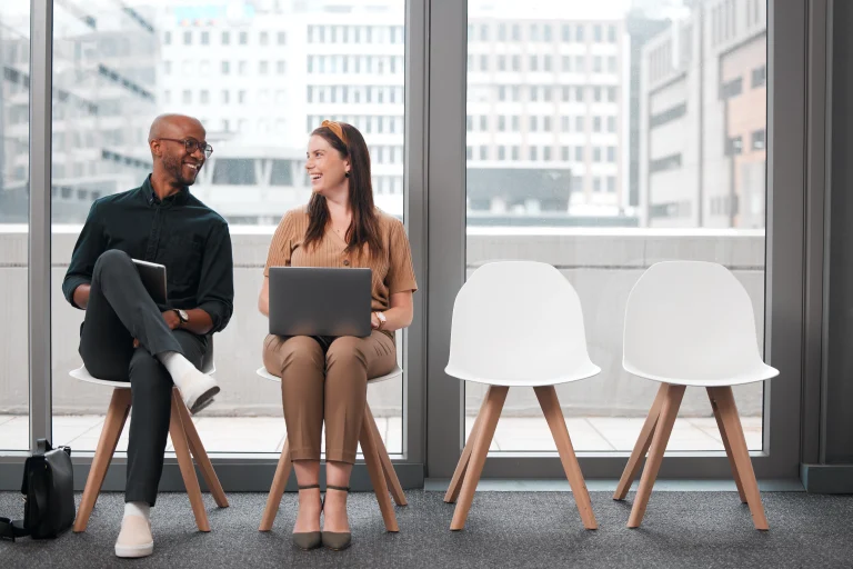 Twee collega's zitten naast elkaar en lachen naar elkaar. Er staan twee lege stoelen naast. Het lijkt op de verpersoonlijking van Blockchain vs Peppol.