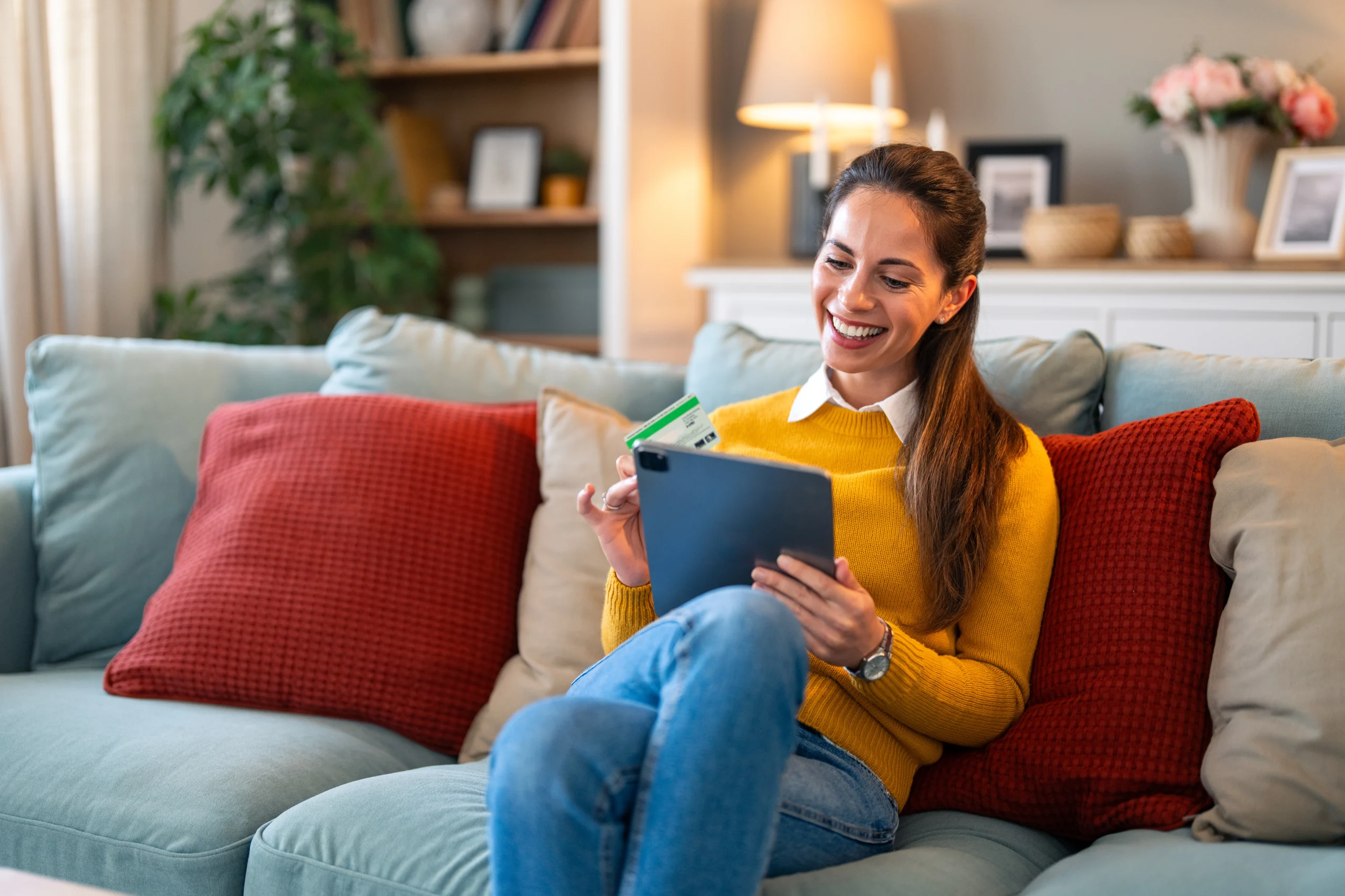 Een vrouw zit lachend met haar tablet op de bank om een automatische incasso in te stellen waarmee ze veel tijd zal besparen.